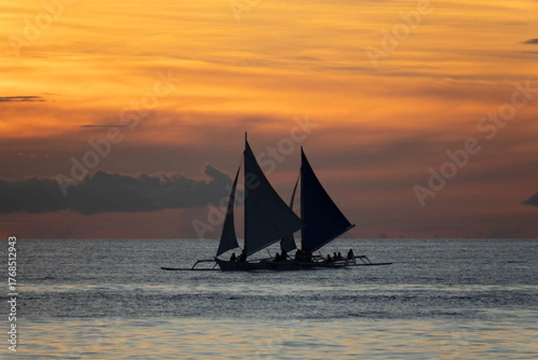 Fototapeta Silhouetted sailboat gliding on calm sea at sunset with vivid orange and golden sky