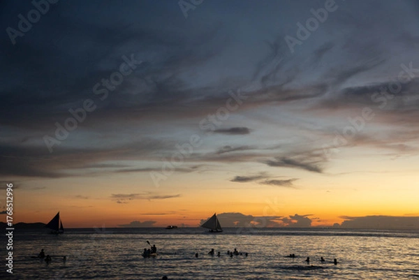 Fototapeta People swimming and sailboats cruising on calm sea during golden sunset with dramatic clouds