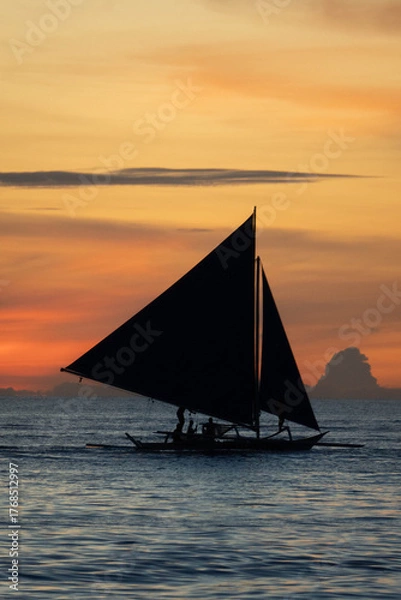 Fototapeta Silhouette of a sailboat gliding over calm sea during golden sunset with orange sky and clouds