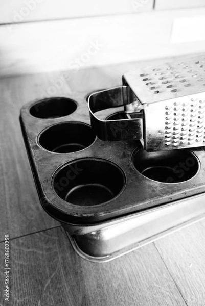 Obraz Black and white close-up of essential kitchen tools a classic metal muffin pan and a versatile box grater, ready for baking or culinary tasks, with a rustic, timeless aesthetic
