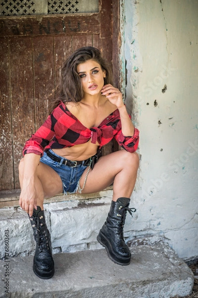 Obraz Young brunette woman with rebellious attitude sits on rustic stone steps, wearing a tied checkered shirt, denim shorts, and boots. Vintage textures and direct gaze evoke bold, rural charm.