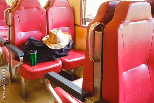 Obraz Traveler’s essentials on red train seat with straw hat, black travel bag, and green tumbler, symbolizing adventure, vacation, and journey lifestyle on public transportation or rail travel, side view