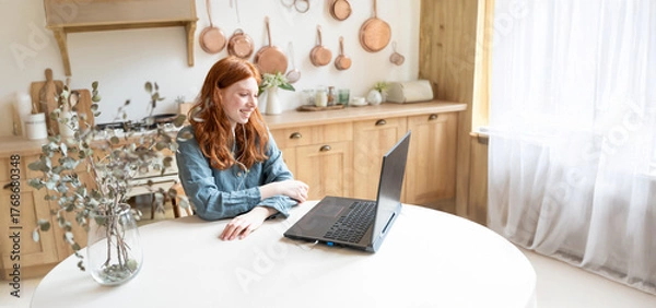 Fototapeta beautiful smiling red-haired girl sits at a table in a forge and has an online video call conference with a laptop. looks at the screen and smiles. remote work, communication at a distance