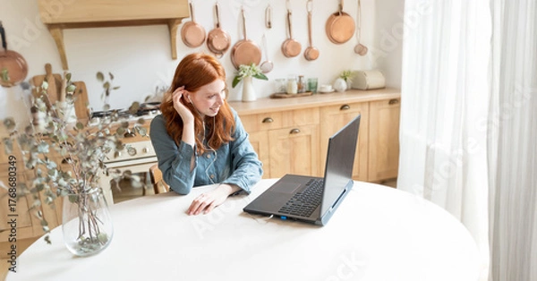 Fototapeta beautiful smiling red-haired girl sits at a table in a forge and has an online video call conference with a laptop. looks at the screen and touches her hair. remote work, communication at a distance