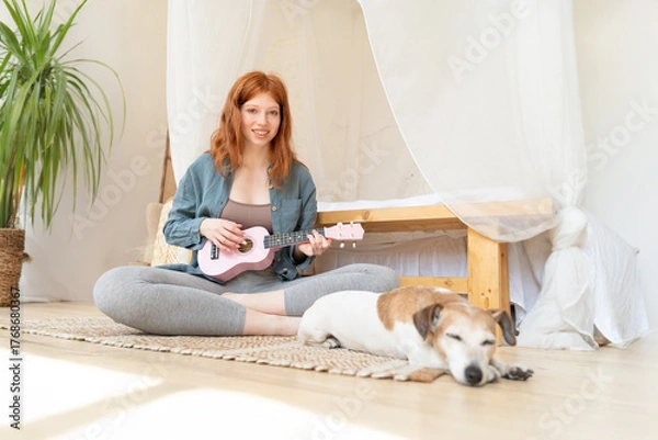 Fototapeta happy red-haired girl in blue shirt plays pink ukulele while sitting on floor in her sunny cozy bedroom. small dog Jack Russell terrier is sleeping next to her. Relaxed home hobby creativity time
