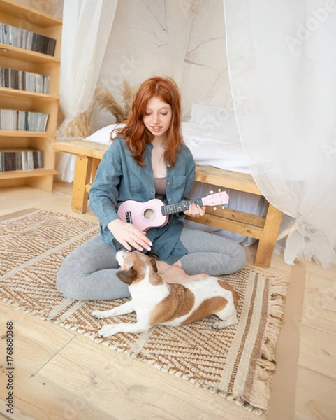 Fototapeta red-haired girl in blue shirt plays pink ukulele while sitting on floor in her sunny cozy bedroom. small dog Jack Russell terrier is watching her attentively. Relaxed home hobby creativity time