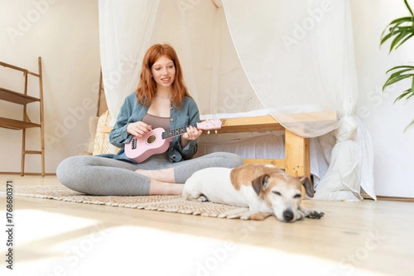 Fototapeta Young red-haired girl in blue shirt plays pink ukulele while sitting on floor in sunny cozy bedroom. small dog Jack Russell terrier is sleeping next to her. Relaxed home hobby creativity time