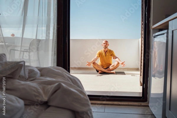 Fototapeta Middle-aged man doing morning yoga breathing exercises in Sukhasana easy pose on sunny summer terrace bedroom. Relaxed lifestyle concept with healthy routine, calm body and mind relaxing.