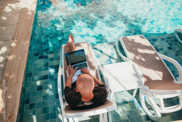 Fototapeta Top view of bald man freelancer CEO business owner working laptop on sun lounger by luxury hotel swimming pool. Digital nomad successfully managing company remote tropical vacation sunlit summer day.