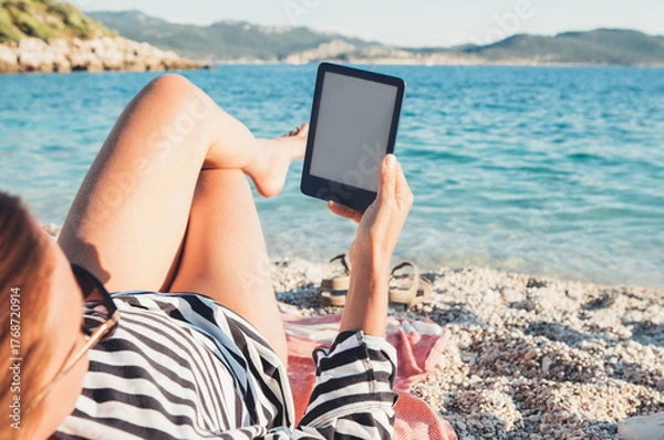Fototapeta Relaxed woman lying on pebble beach reading e-book on digital tablet during sunny summer day. Concept of leisure, technology, travel, slow life vacation near turquoise sea water with coastal mountains