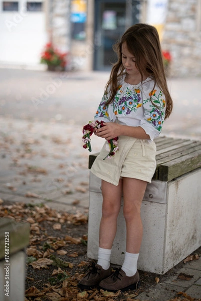 Obraz Sad Ukrainian girl sitting outdoors and leaning on a wooden bench while looking thoughtfully at a floral wreath in her hands among yellow autumn leaves, concept of emotion and Ukrainian tradition