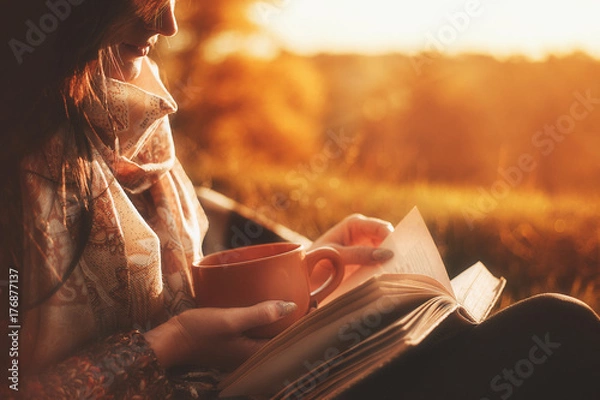 Fototapeta a woman sits near a tree in an autumn park and holds a book and a cup with a hot drink in her hands. Girl reading a book