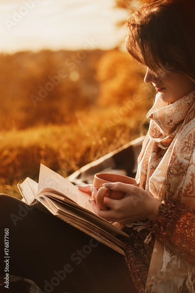 Fototapeta a woman sits near a tree in an autumn park and holds a book and a cup with a hot drink in her hands. Girl reading a book