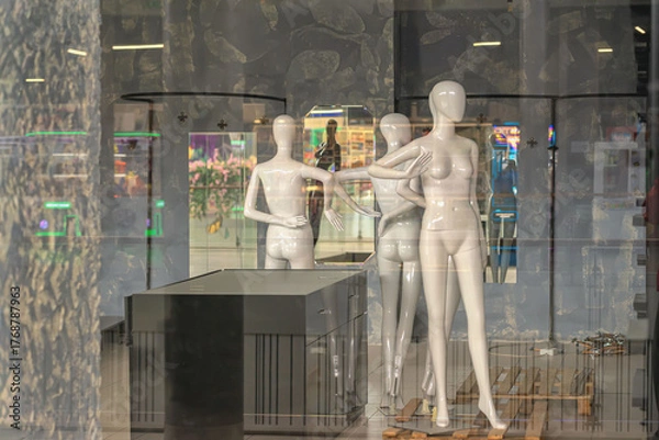 Fototapeta Mannequins displayed in shop window with reflections of mall interior and empty retail setup. Concept of fashion retail, storefront visuals, display design