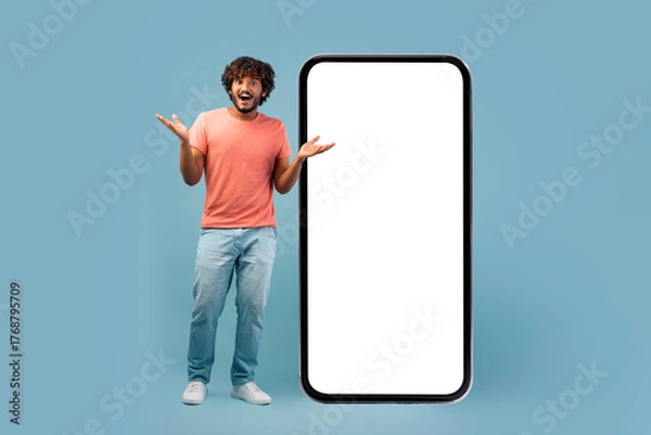 Obraz Cool eastern man displays a surprised expression while standing next to a large smartphone with a white screen. He gestures enthusiastically, showcasing a fantastic online opportunity.