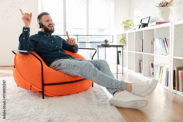Fototapeta A man listens to music through headphones while sitting comfortably in an orange bean bag chair. Sunlight fills the room, highlighting books and plants around him.