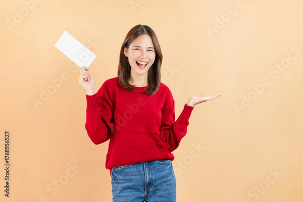 Fototapeta Happy Asian business woman standing and hold blank paper or gift voucher mockup on beige color background.