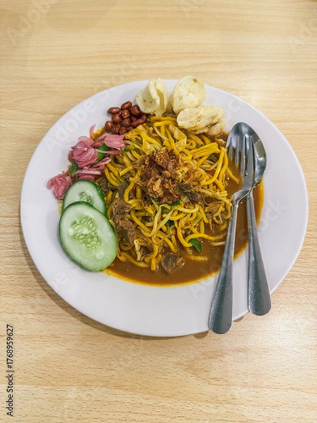 Fototapeta A plate of traditional Indonesian Mie Aceh served with rich spicy curry sauce, beef, cucumber slices, peanuts, crackers, and fried shallots on top.