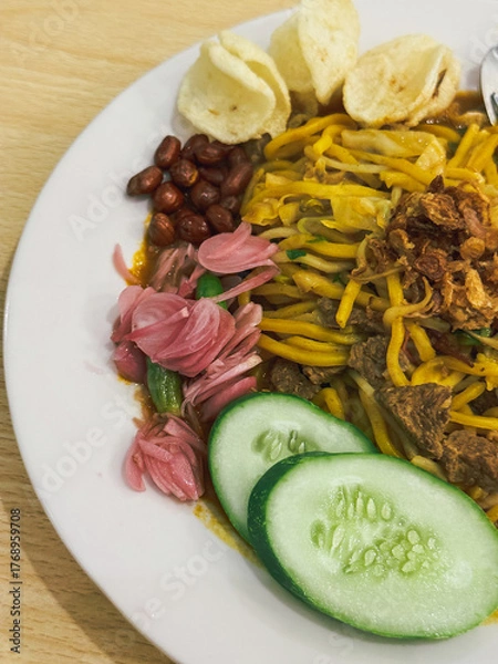 Fototapeta A plate of traditional Indonesian Mie Aceh served with rich spicy curry sauce, beef, cucumber slices, peanuts, crackers, and fried shallots on top.