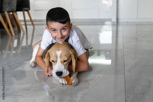 Fototapeta A young boy smiles while resting on a tiled floor beside his beagle. The dog focuses on chewing a tasty treat, showcasing their friendship in a cozy home setting.