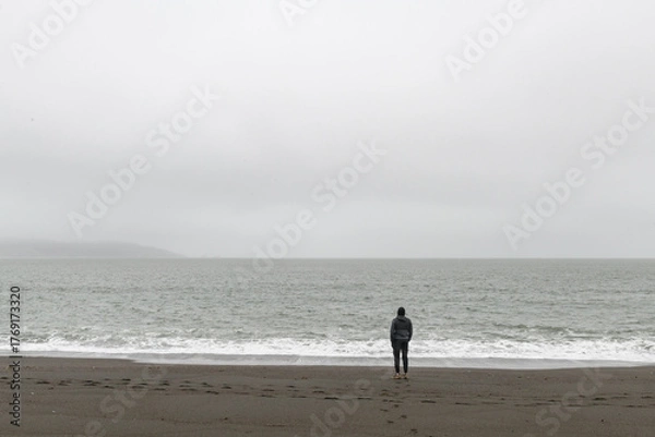 Fototapeta A lone individual man stands on a isolated beach, back turned and looking out in contemplation at a vast fog-enshrouded ocean.