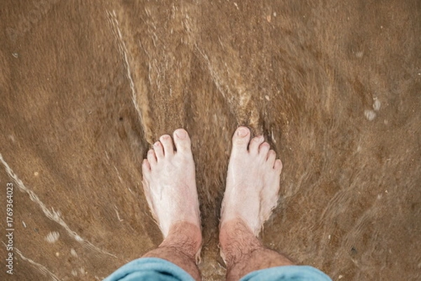 Fototapeta Caucasian male bare feet and pulled up jeans, standing on a sandy beach with shallow tidal waves sea water reaching the shore. Top down, close up view