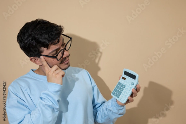 Obraz Man with glasses looking at a blue calculator with a thoughtful expression on a light brown background with copy space, concept of financial analysis and economic planning