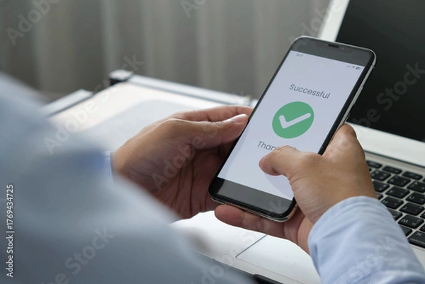 Fototapeta Successful mobile transaction with green checkmark displayed on smartphone screen held by man in office setting