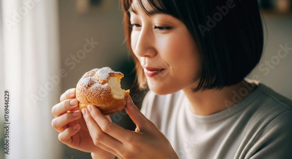 Obraz Happy Japanese woman savoring a sweet cream puff, her expression showing pure delight while enjoying a delicious dessert.