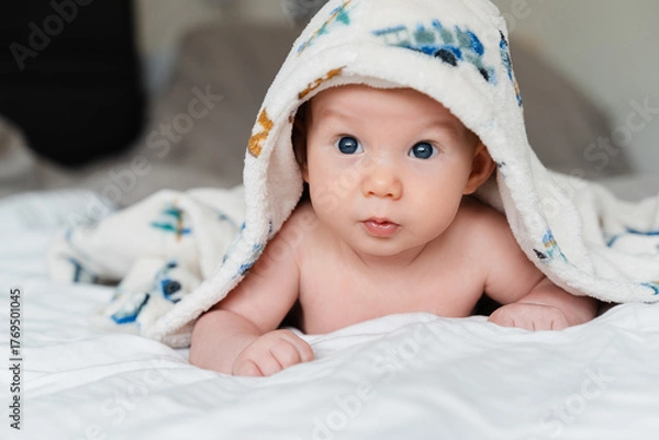 Fototapeta Happy curious baby child after bath with towel on head. Adorable little boy wrapped in blanket. Cutest 3 months old baby smiling in towel on bed close up at home. Child caring routine.