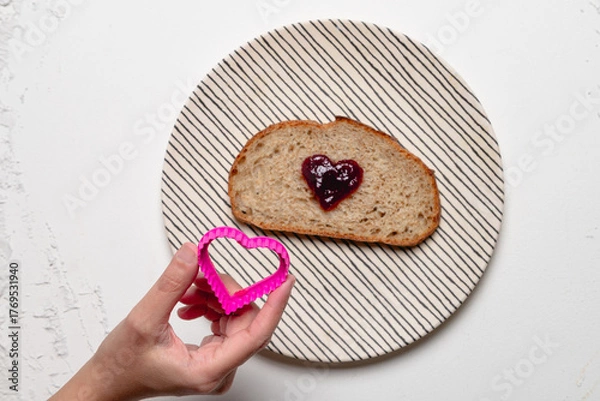 Fototapeta A hand holding a pink heart-shaped cookie cutter next to a slice of bread with heart-shaped jelly on a striped plate, over a white background.