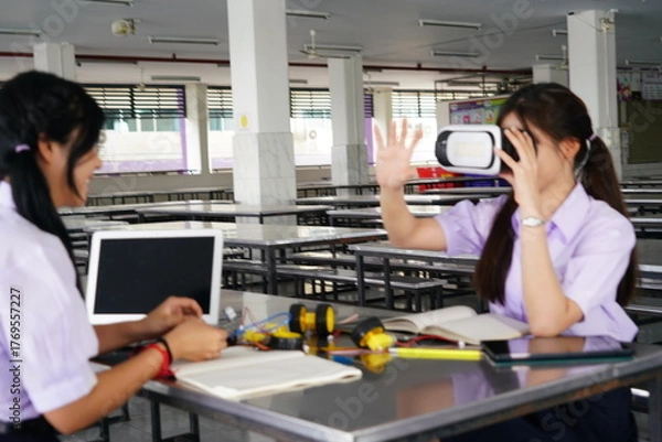 Fototapeta  teenage female student in her school uniform sits in class doing homework and working on a robot circuit with her female friend wearing VR glasses in the cafeteria at a high school.