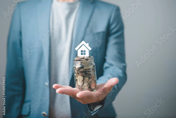 Fototapeta Businessman holding a jar full of coins with a house icon above it, symbolizing real estate investment, home savings, financial planning, mortgage, and property investment concept.