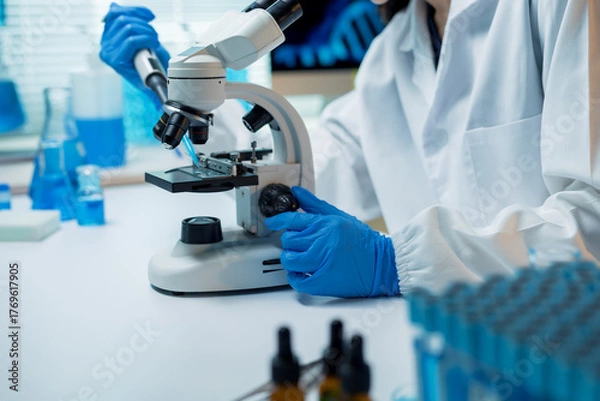 Fototapeta Scientist wearing a lab coat and blue gloves performing an experiment with a microscope and pipette, focusing on biology and genetics