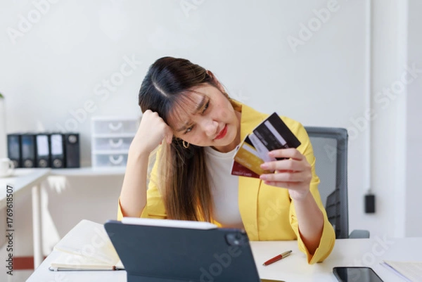 Fototapeta Asian woman feeling worried and stressed, sitting at office desk, looking at credit cards, dealing with overspending debt