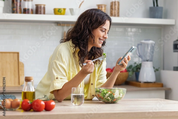 Fototapeta Beautiful woman eating healthy salad while using smartphone in the kitchen at home