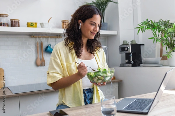Obraz Beautiful woman with earphones eating salad while using laptop in the kitchen at home