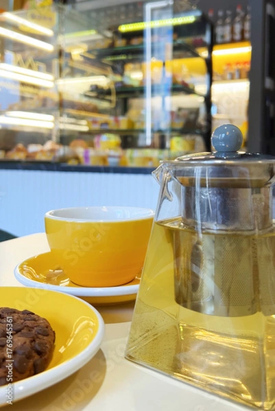 Fototapeta A yellow cup and teapot with tea are on the cafe table. Blurred background of the cafe interior.Vertical 