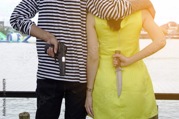 Fototapeta Cheating couple, a man put his arm around woman shoulder looking at the lake in the park. Boyfriend holding a gun girlfriend holding a knife in behind