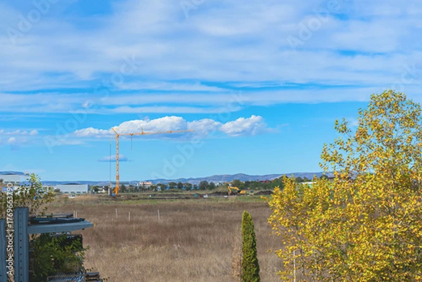 Fototapeta Yellow tower crane working on open construction site in rural field with distant buildings. Concept of land development, suburban expansion, construction progress, urban sprawl