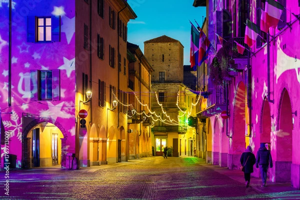 Fototapeta Small Italian town of Alba at dusk, with narrow cobblestone streets lined with historic buildings and illuminated by Christmas lights.