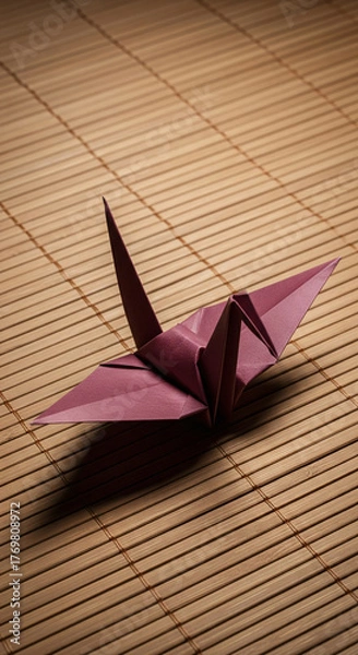 Fototapeta Traditional Japanese origami crane in a rich burgundy color resting on a bamboo mat. A serene still life representing peace, hope, and the delicate art of paper folding, with dramatic lighting.
