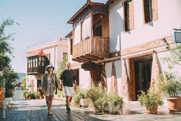 Fototapeta Middle aged couple walking hand in hand along narrow street in Kas town historical center. Romantic travel, love relationship, honeymoon vacation and happy retirement lifestyle concept.