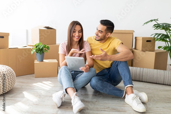 Fototapeta A happy couple sits on the floor in their new home, sharing a moment while looking at a digital device. They are surrounded by moving boxes, suggesting they are settling in.