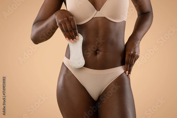 Fototapeta Unrecognizable black woman poses in underwear while holding a menstrual pad against a beige background. This showcases an important aspect of female hygiene and comfort during menstruation.