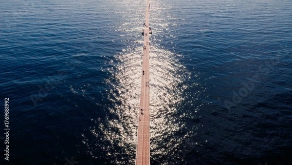 Obraz People walk along a wooden pier over calm water reflecting sunlight on a clear day