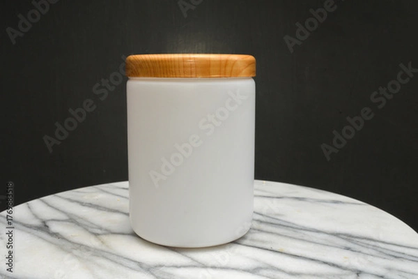 Obraz mockup of a white frosted jar, bottle with a wooden lid, sitting on a white marble table with a dark background. Ideal for beauty, cosmetic, food, and packaging design.