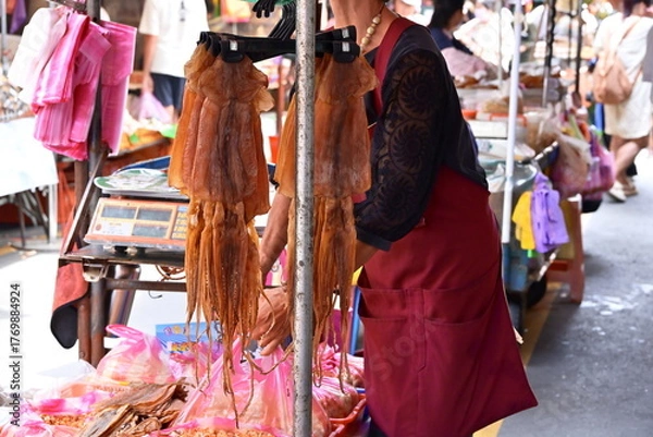 Fototapeta Taiwan - Sep. 20, 2025: Market vendor in black shirt and red apron behind stall with hanging dried brown squid, packaged goods in plastic bags on table, red scale visible, busy market background.
