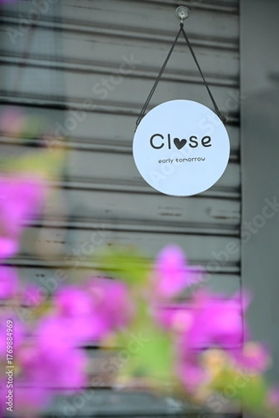 Fototapeta Taiwan - Sep. 20, 2025: White round sign with "Close early tomorrow" and heart on gray shutter, blurred pink-purple bougainvillea in foreground, selective focus composition, Taiwan storefront.