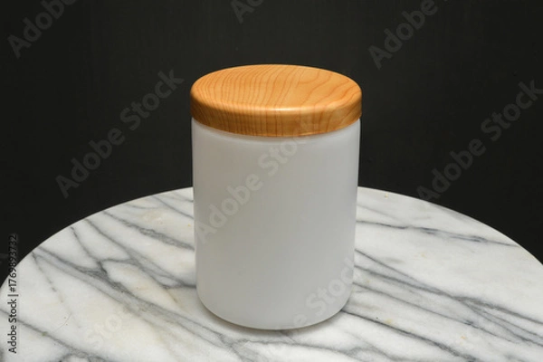 Fototapeta mockup of a white frosted jar, bottle with a wooden lid, sitting on a white marble table with a dark background. Ideal for beauty, cosmetic, food, and packaging design.
